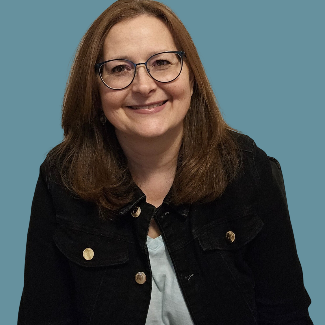 Une femme souriante avec des lunettes, portant une veste noire, assise devant un fond bleu.